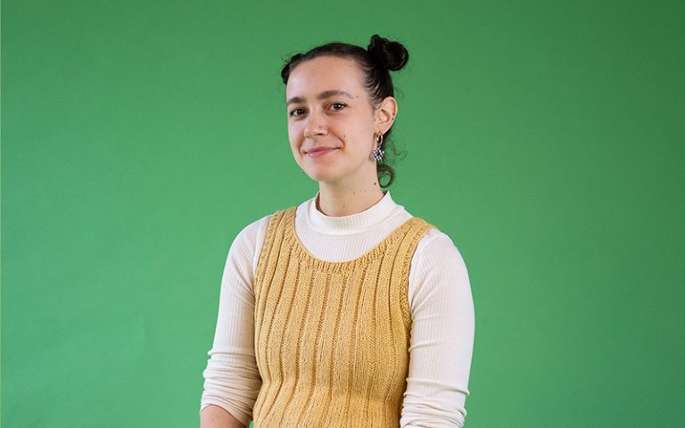 Photograph of student Indianna Williams. She is wearing a yellow knitted top over a white shirt. She it sitting in front of a green backdrop.