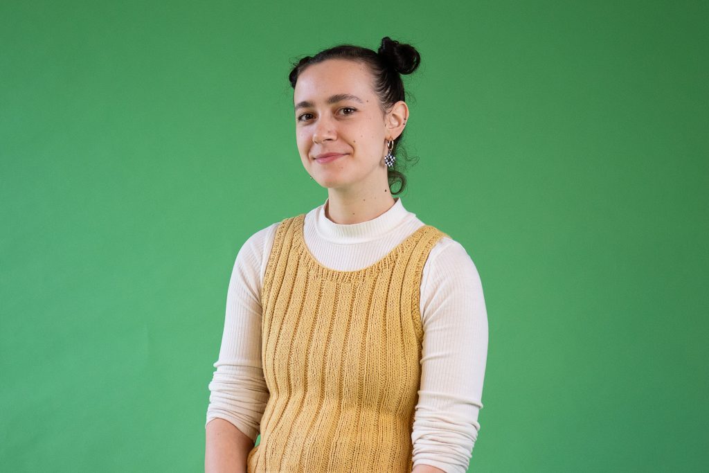 Photograph of student Indianna Williams. She is wearing a yellow knitted top over a white shirt. She it sitting in front of a green backdrop.