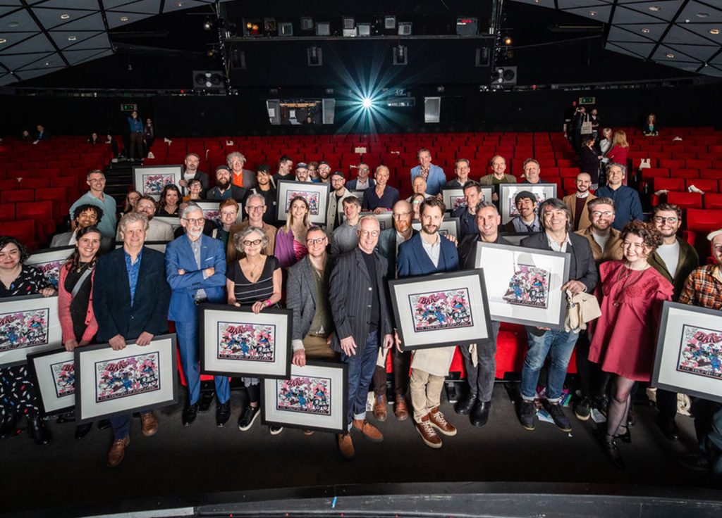 Group photo of the BAA award winners. Everyone is standing in at the front of a theatre seating; a projector is lit up at the back of the room.