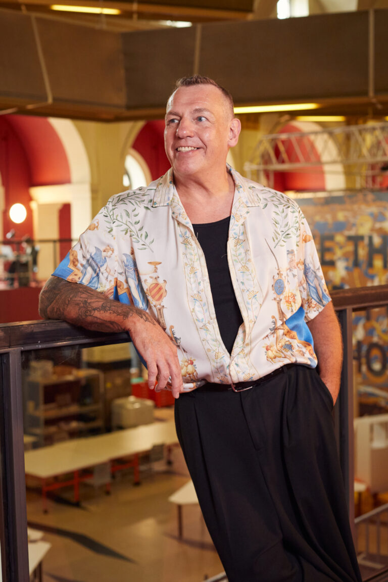 A person stands on an indoor balcony, smiling and looking to the side. They are wearing a patterned short-sleeved shirt over a black top with black pants. The background includes red and white walls and various tables and equipment.