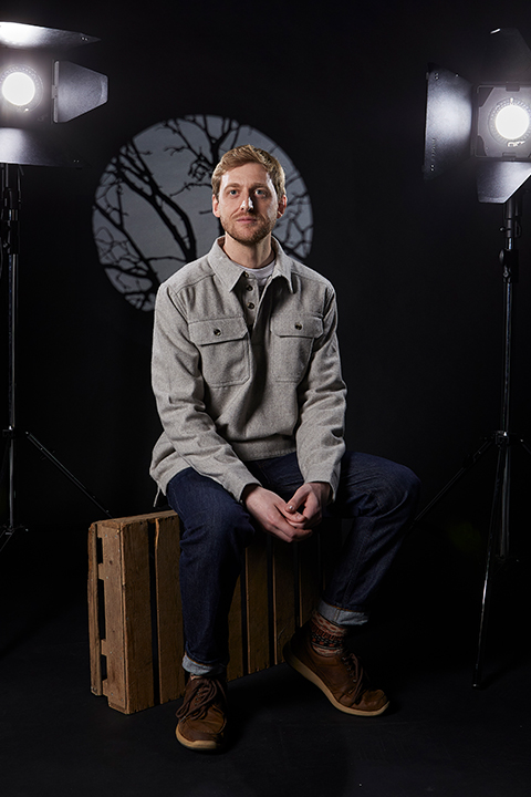 A man sits on a wooden crate in a dimly lit studio. He's wearing a gray jacket and jeans, and is surrounded by studio lights. A circular image of tree branches is visible in the background.