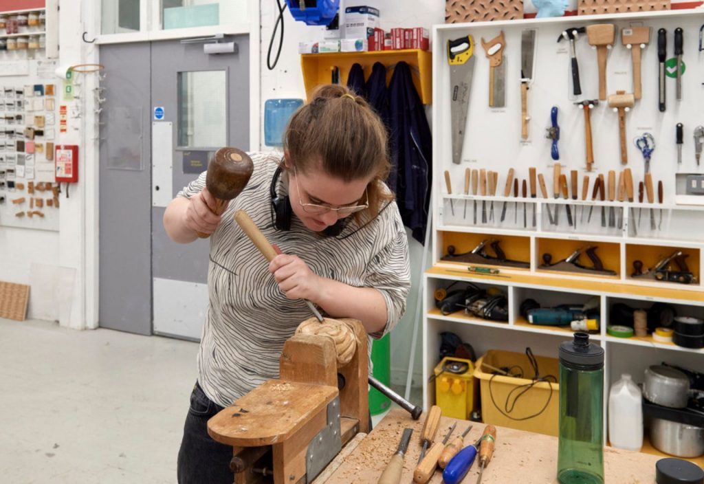 A Fine Art student chiseling their project which is in a wooden clamp in the 3D studio. They have their hair up and googles on. In the background, there is lots of equipment and hardware hung on the wall.