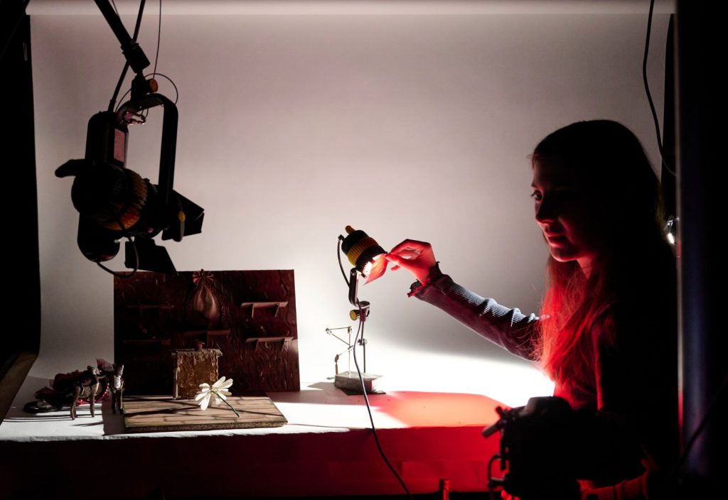 A student holding up a red gel to a light in the animation studio, they are shooting a scene. They have long hair. In the foreground is a camera hanging from top left hand corner.