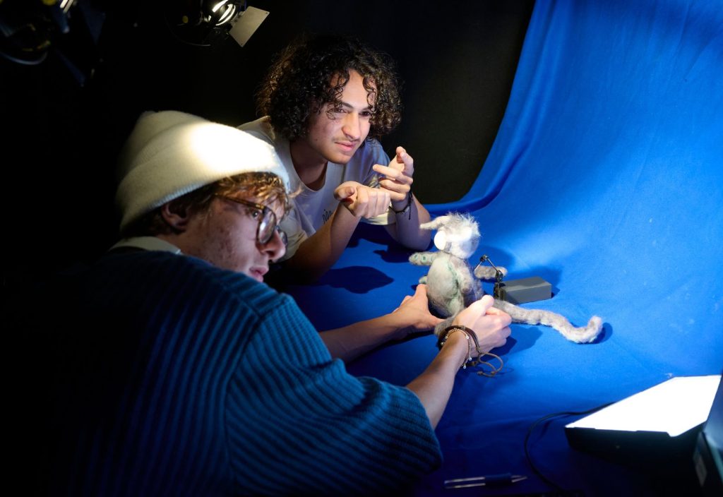 Two students lean in to shoot their project in the animation studio. They are holding a character on a blue background. One has a white hat on and glasses, the other has curly dark hair and is wearing a t-shirt.