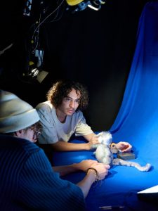 Two students lean in to shoot their project in the animation studio. They are holding a character on a blue background. One has a white hat on and glasses, the other has curly dark hair and is wearing a t-shirt.