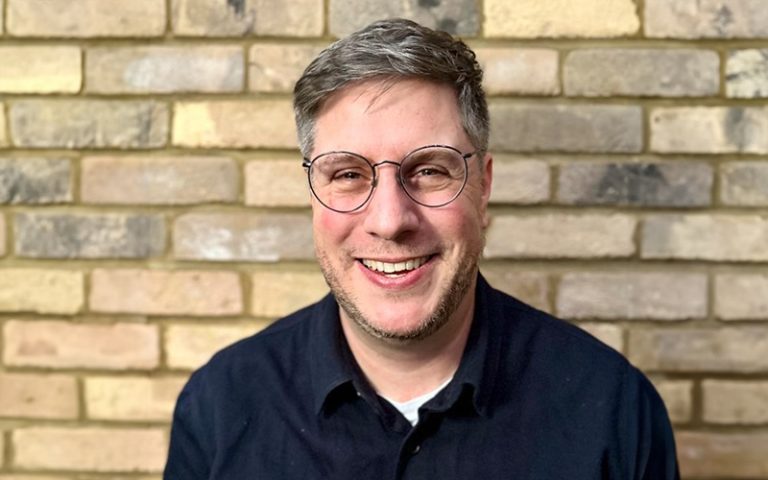 Photo of Ben Stopher. Ben has glasses and a navy shirt, in front of a brick wall.