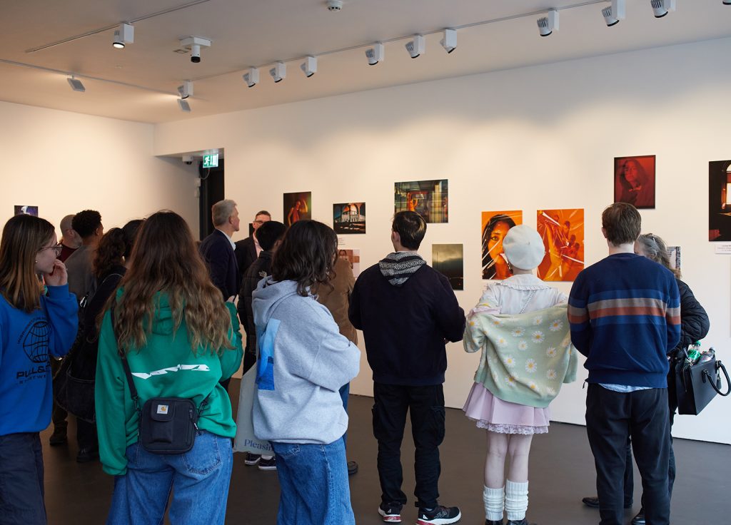 Photograph of a crowd of people in a gallery; they mostly have their backs to the camera, looking at photographs on the gallery wall.