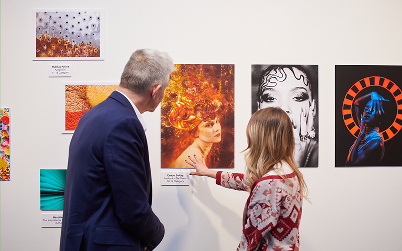 Two people are looking at photographs on a gallery wall. One is pointing ay one of the images.