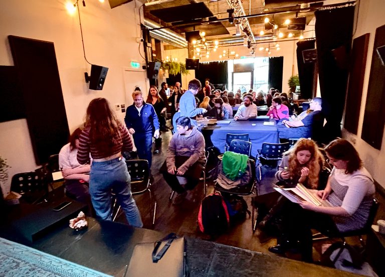 A large room full of people. People are gathering around several tables with blue table cloths.