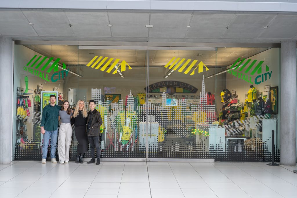 A photograph of BA (Hons) Interior Design students stood in-front of a glass shop window. The shop window was designed by the students for a local business called 'On the stool city'.