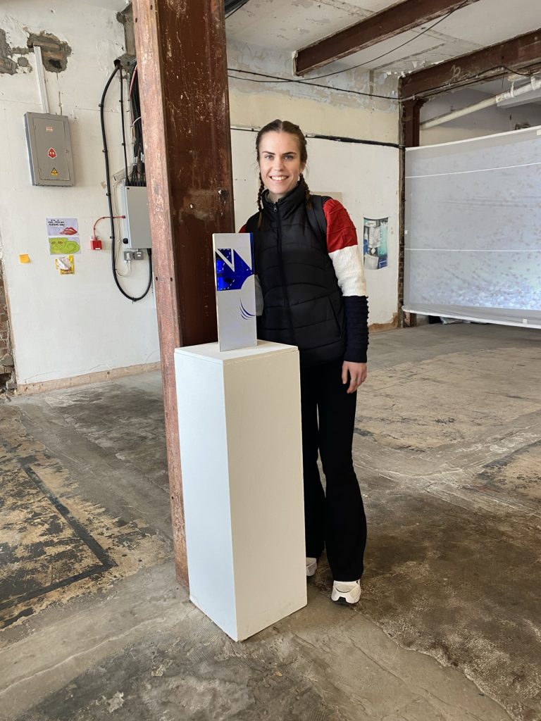 A person with long hair, dressed in a black jacket, stands indoors next to a tall white pedestal displaying a small rectangular art piece. The room has exposed beams and industrial elements.