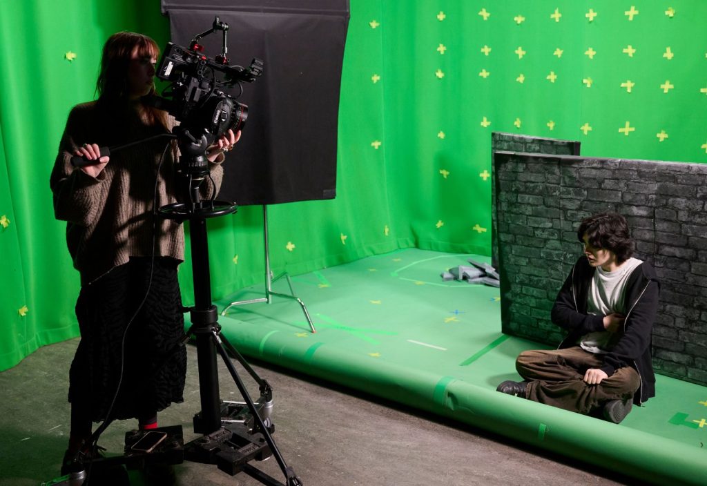 Two student shooting a project. One is sitting cross-legged on the floor in front of a prop wall, the other is at a camera recording. Background is green screen.