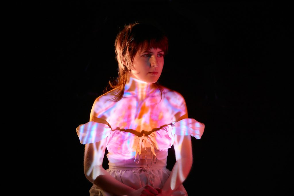 A woman in a white blouse illuminated by colourful light projections creating abstract patterns on her torso and face, set against a dark background.