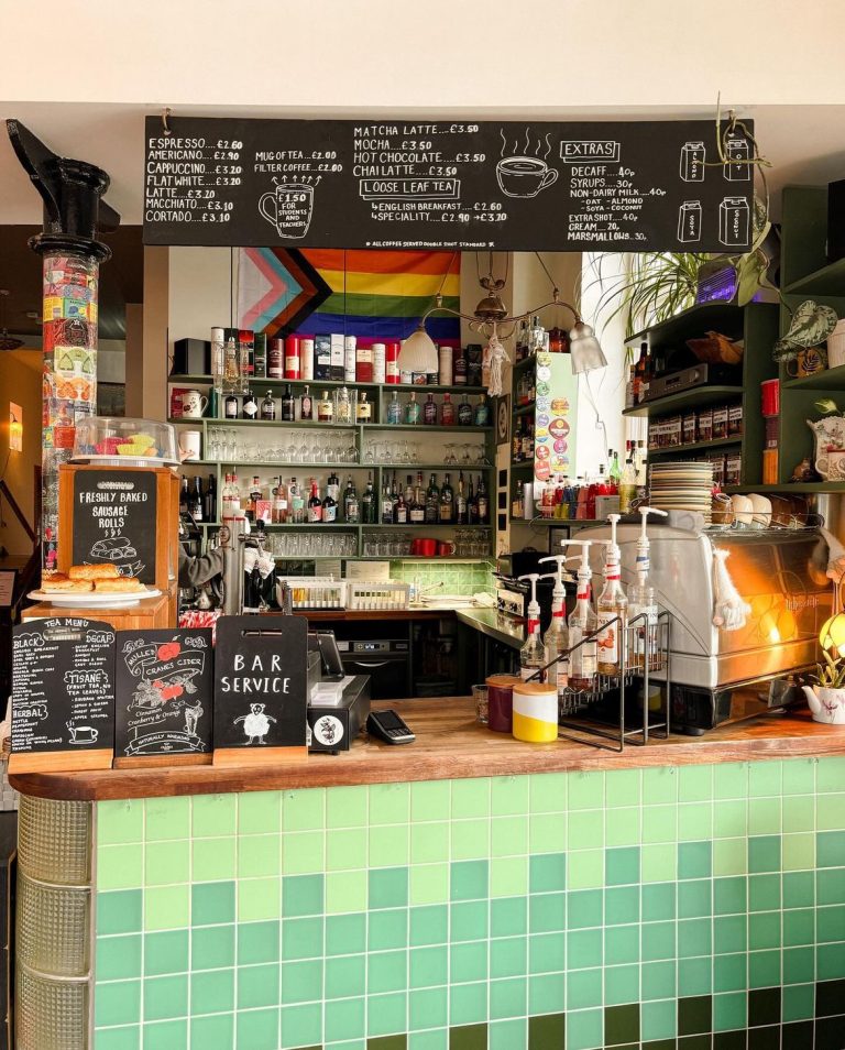 Photograph of the Merchants House bar. A rainbow flag is hung behind the bar, above stacked bottles and jars. Various chalkboards with the menus are hung around the bar.