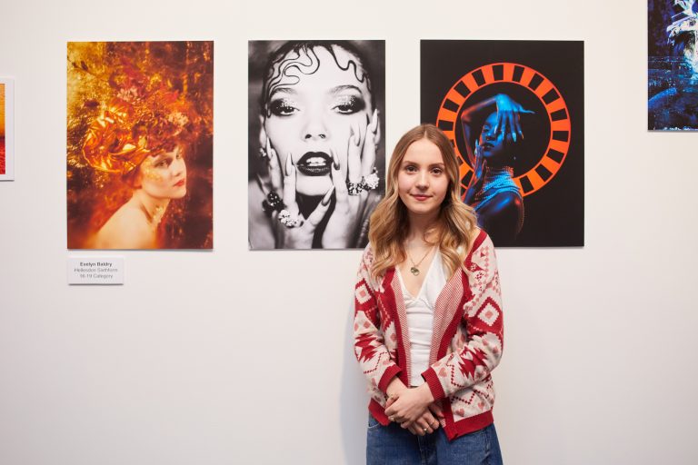 Photograph of a person in a white t shirt and pink cardigan. She stands in front of three photos.