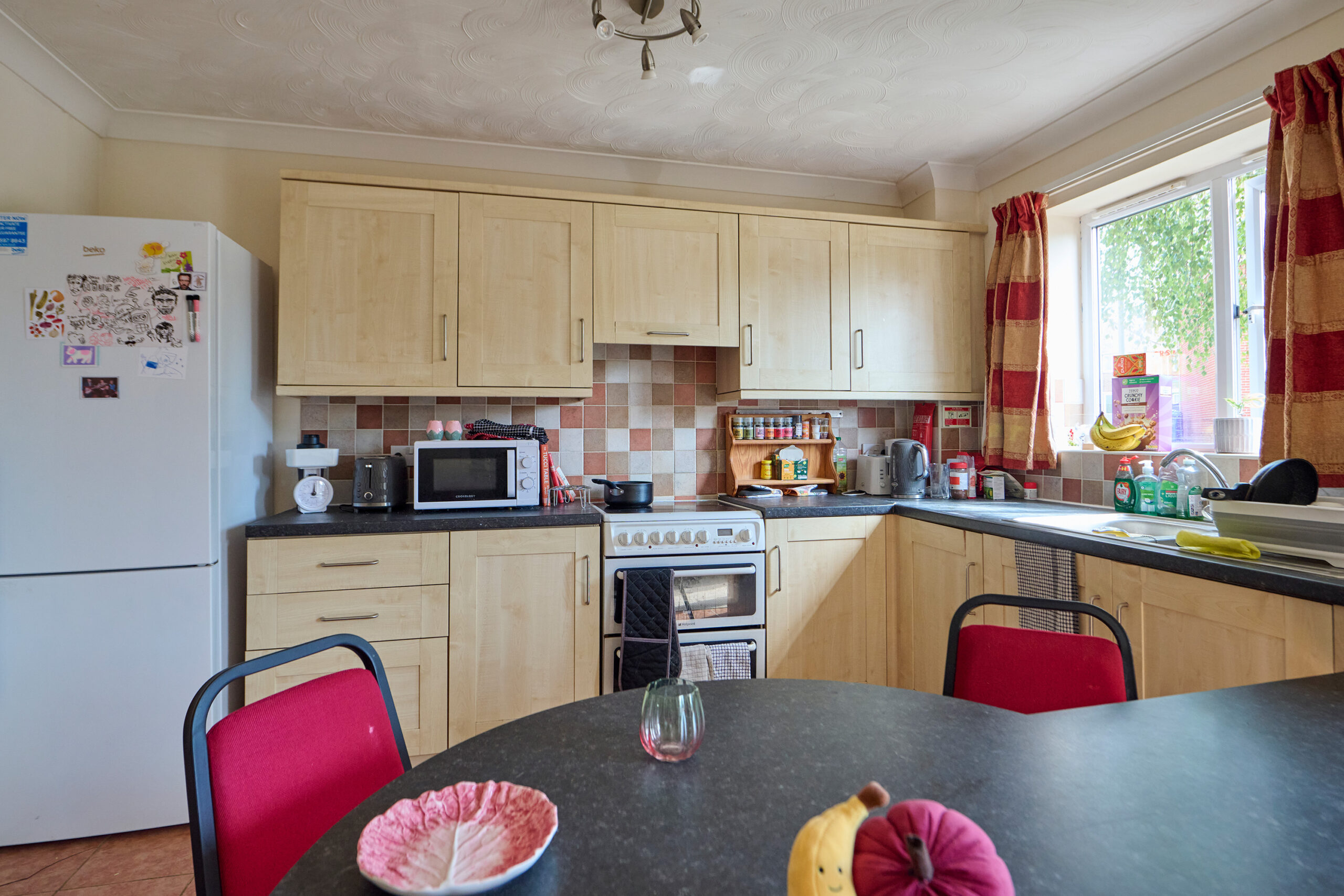 A cozy kitchen with wooden cabinets, a white fridge, microwave, stove, and various small appliances. A round black table with red chairs is in the foreground. Sunlight comes through a window with red curtains.