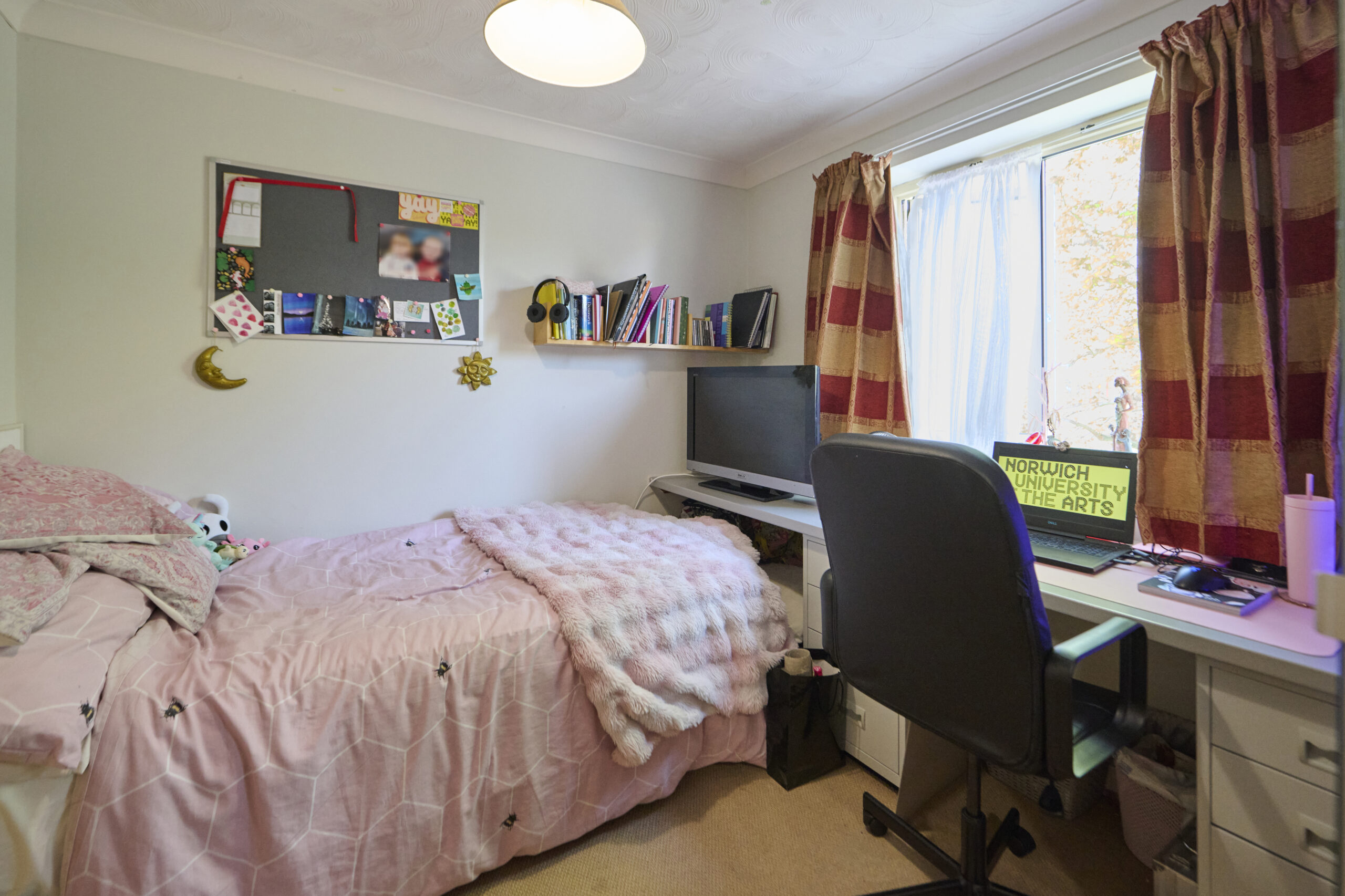 A cozy bedroom with a single bed covered in a pink blanket, a desk with a computer, books, a chair, a window with red and beige curtains, and a bulletin board with photos and notes on the wall.