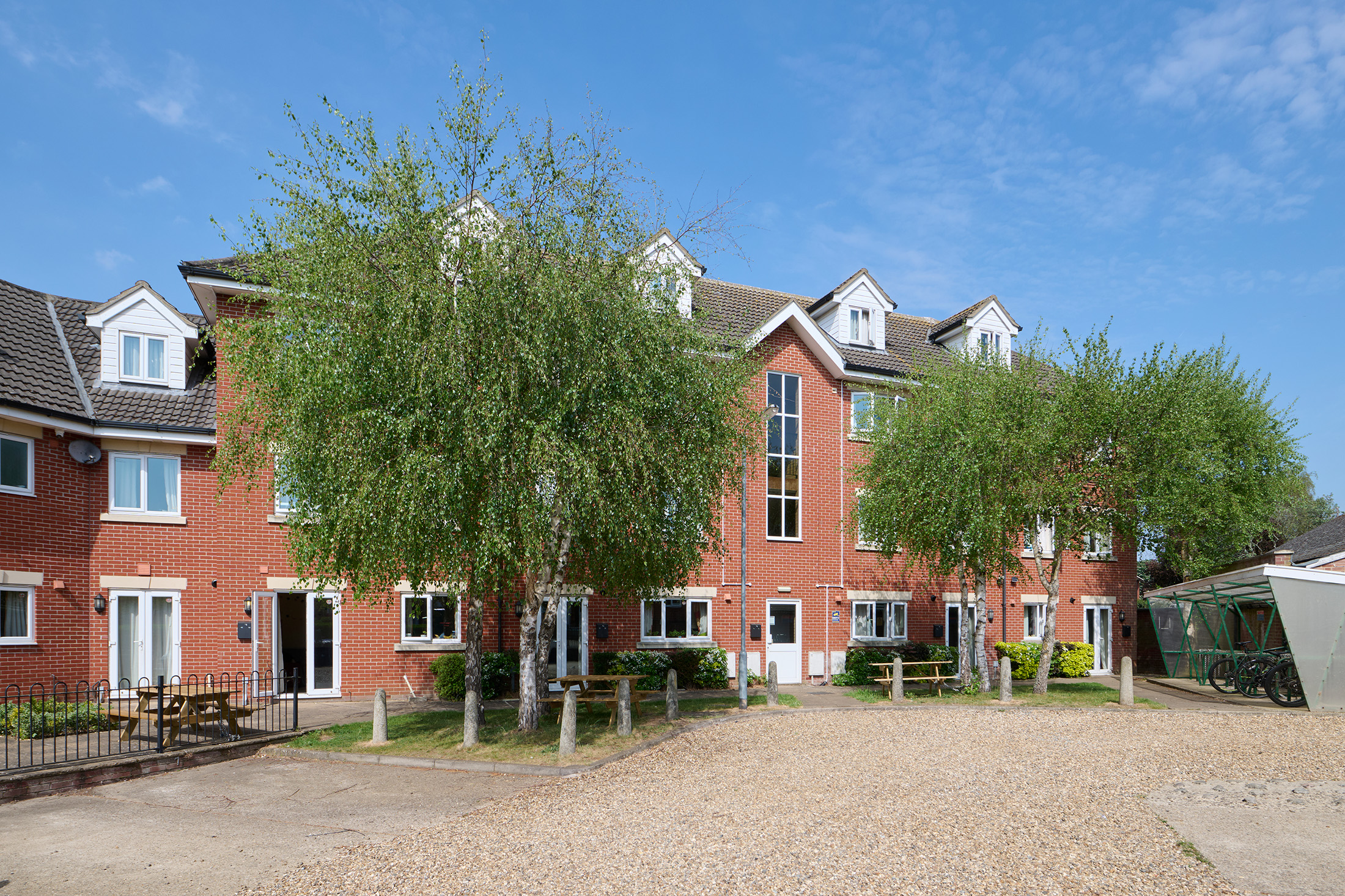 A modern red-brick apartment building with multiple floors and dormer windows, surrounded by trees and a gravel parking area under a clear blue sky.