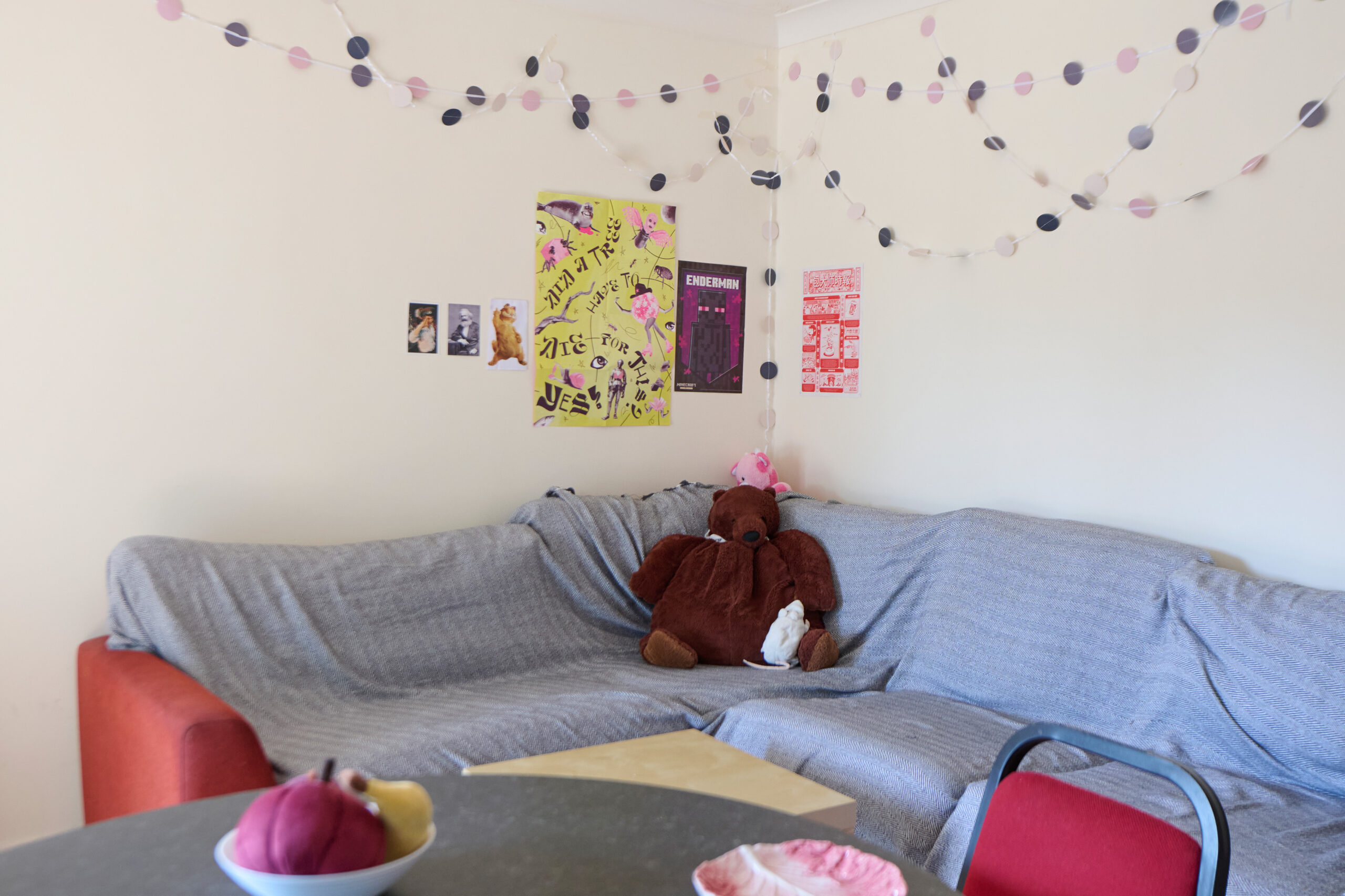 A cozy living room features a red and gray sectional sofa covered with a gray sheet, a large teddy bear, a stuffed toy, string garlands on the wall, colorful posters, and a table with a fruit bowl in the foreground.