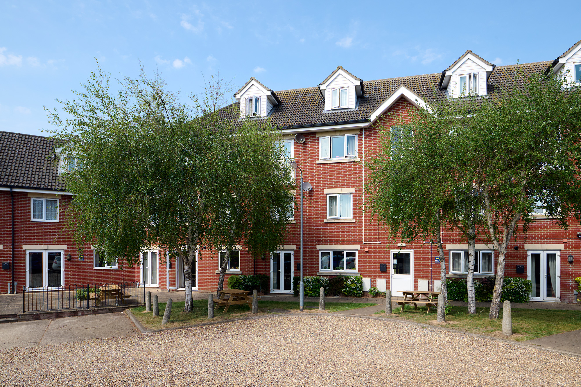 A modern red-brick apartment building with white trim and dormer windows, surrounded by small trees, benches, and a gravel courtyard under a blue sky.