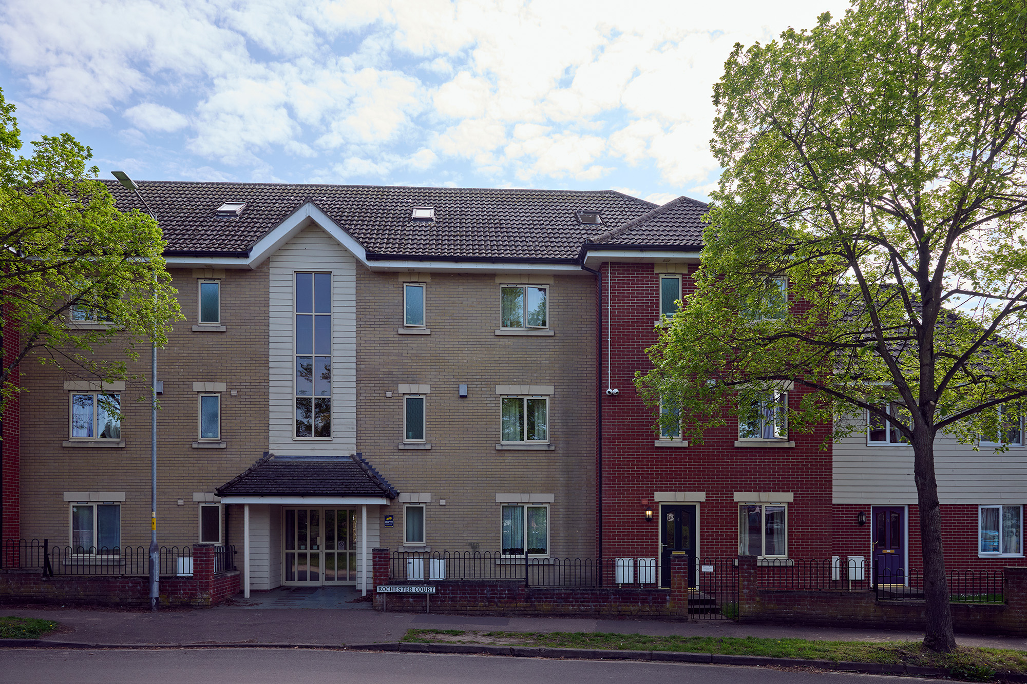 A modern, three-story apartment building with brick and light-colored siding, large windows, and a central glass entrance. Trees with green leaves line the street in front under a partly cloudy sky.