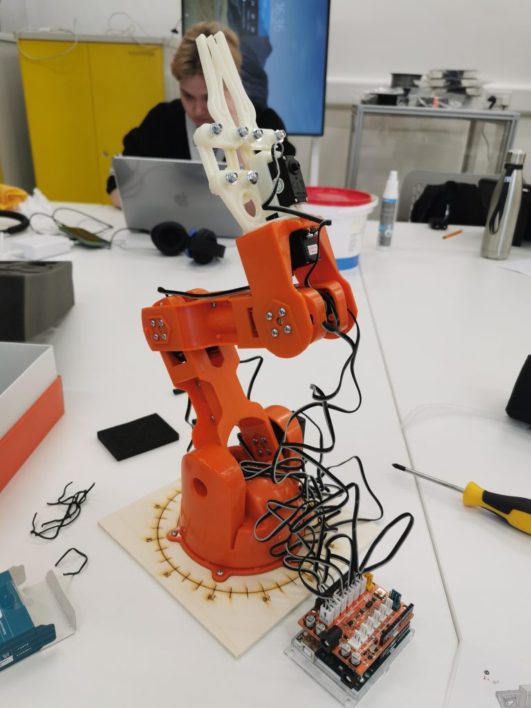 An orange robotic arm on a worktable, surrounded by electronic components and wires, in a busy workshop environment.