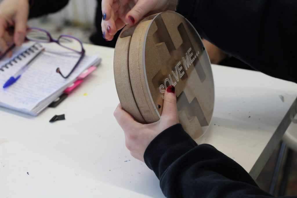 A person wearing a black sweater holds a circular wooden puzzle with the word "SOLVE ME" engraved on it. Another person is visible in the background, writing in a notebook with glasses placed on the table nearby.