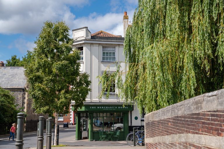 A photograph of The Merchants House in Norwich. The image was taken in the summer on a bright and sunny day. The photo is of the coffee shop entrance from a short distance away with various greenery from surrounding tress falling into frame.