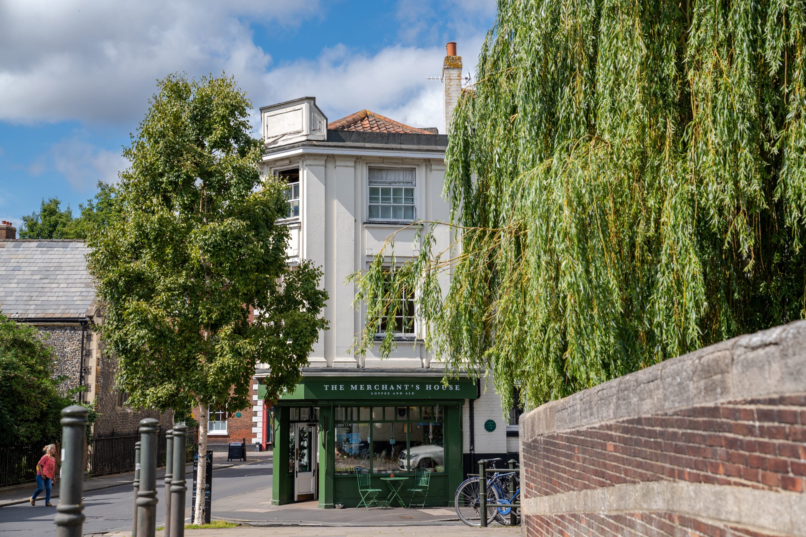 A photograph of The Merchants House in Norwich. The image was taken in the summer on a bright and sunny day. The photo is of the coffee shop entrance from a short distance away with various greenery from surrounding tress falling into frame.