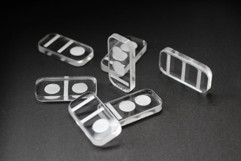 Several rectangular transparent acrylic dominos-like sculptures with different geometric patterns, including circles and lines, are scattered on a solid dark surface. Some pieces are laying flat, while one stands vertically.