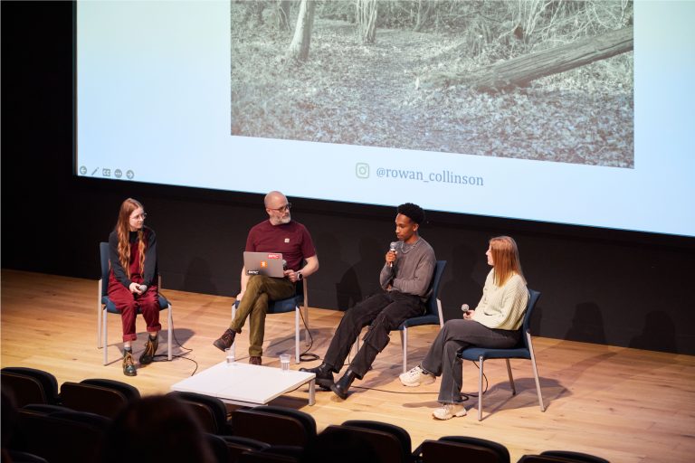 Photograph of four people sitting at the front of a lecture theatre; one person is speaking into a microphone.