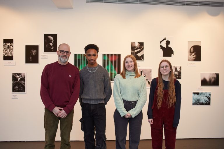 Group photo of four people standing in a line in front of a white gallery wall. A selection of photographs are on the wall behind the group.