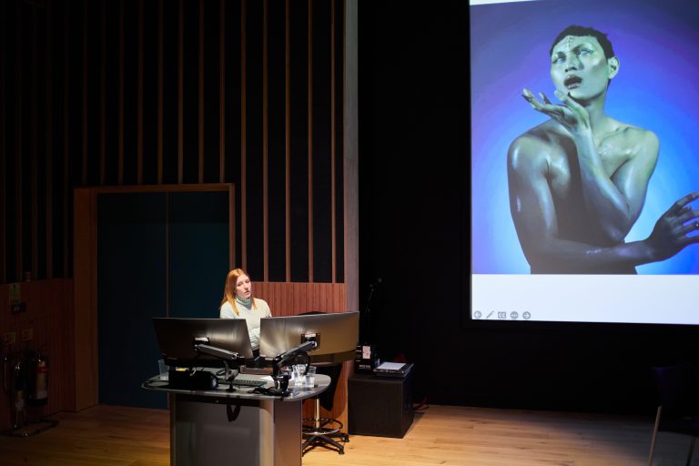 A person is presenting from the front of a lecture theatre; a blue image is on the screen next to them.