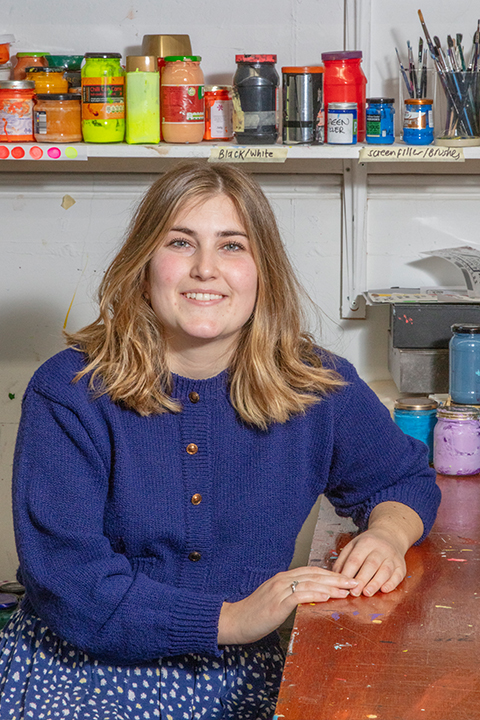 A smiling young woman with shoulder-length hair sitting at a paint-splattered table, surrounded by colourful paint jars and brushes. she wears a blue sweater and spotted skirt.