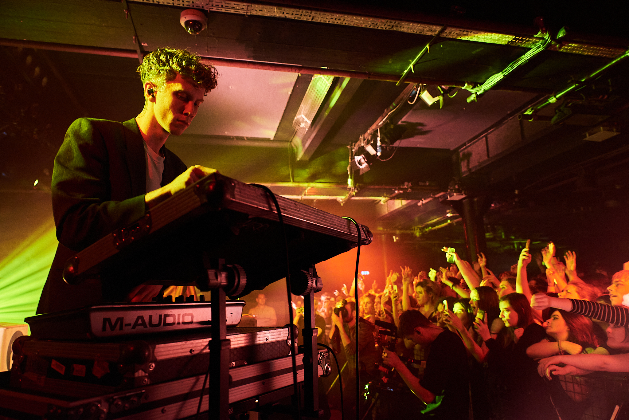 A musician plays an electronic keyboard on stage under colorful lights, while a crowd of energetic fans with raised hands enjoys the live performance in a dimly lit club.