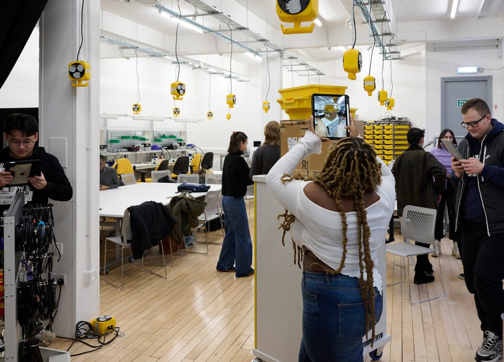 A bustling engineering lab with students engaged in various activities; some discuss and others work on tablets amidst equipment and yellow extension leads overhead.