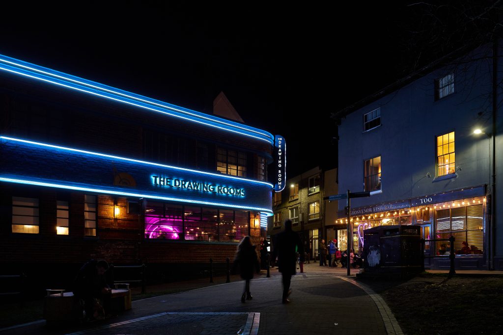 A nighttime street scene with a brightly lit building on the left featuring neon blue lights and a sign that reads "The Drawing Rooms." On the right, people walk past a warmly lit café or restaurant. The street is paved and lined with buildings.