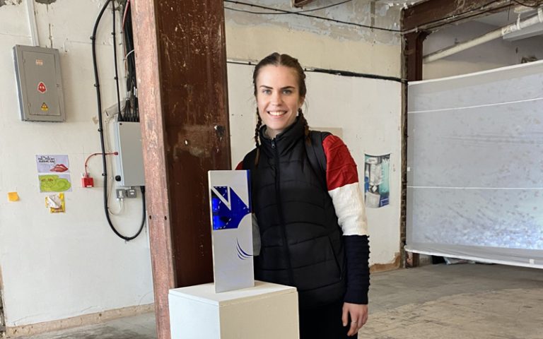 A person with long hair, dressed in a black jacket, stands indoors next to a white pedestal displaying a small rectangular art piece. The room has exposed beams and industrial elements.