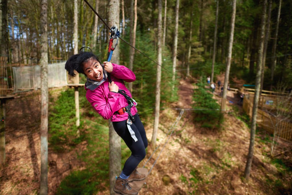 A person wearing a pink jacket is zip-lining through a forest. They appear to be smiling or laughing while holding onto the zip-line harness with both hands. The background shows tall trees and a forest floor covered in leaves.