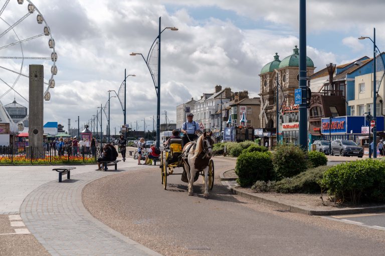 A horse-drawn carriage carrying passengers drives along a seaside promenade with shops, a ferris wheel, and Victorian-style buildings in the background. Pedestrians and cyclists are also visible on the path, and cars are parked along the street.