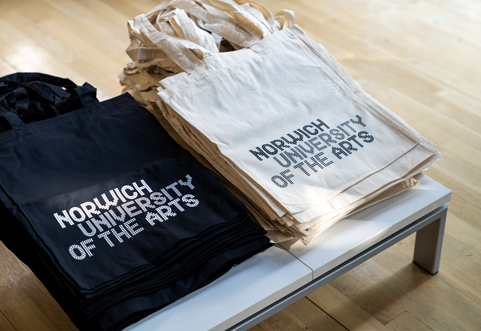 A black and white tote bag with the 'Norwich University of the Arts' logo on the front. The bags are in two separate piles on a table.