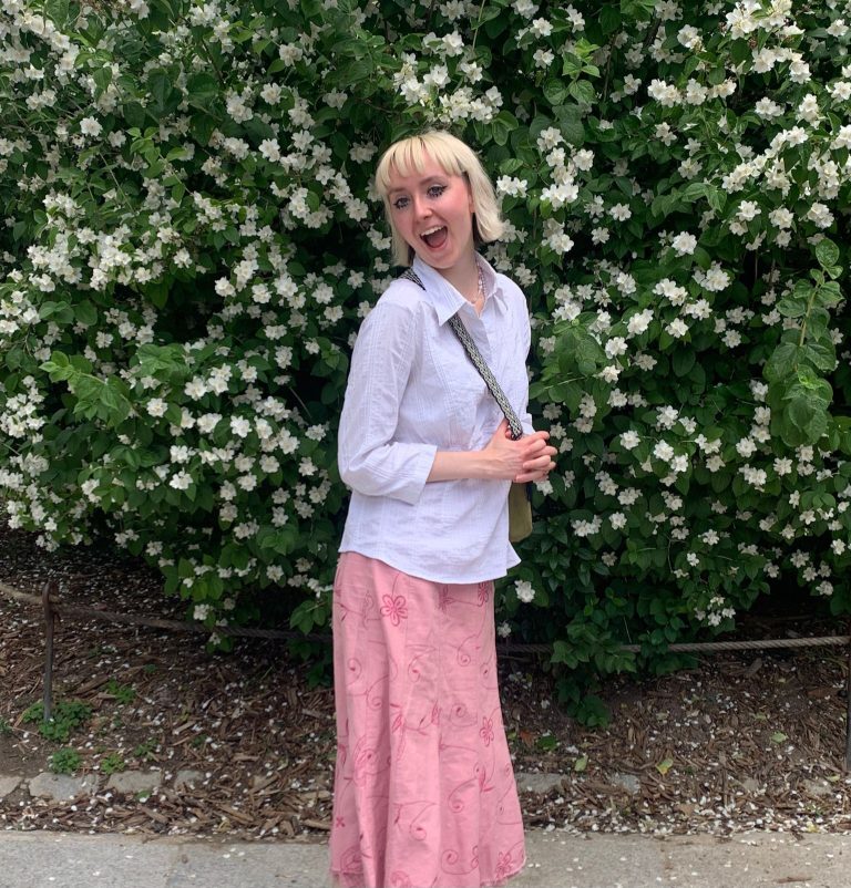 Molly De Smith, a Textile Design student with short blonde hair stands in front of a lush green bush with white flowers, smiling broadly. She is wearing a white shirt and a long pink skirt with a floral pattern. She is holding onto a bag over her shoulder, and her pose exudes happiness and confidence.