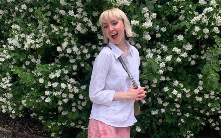 Molly De Smith, a Textile Design student with short blonde hair stands in front of a lush green bush with white flowers, smiling broadly. She is wearing a white shirt and a long pink skirt with a floral pattern. She is holding onto a bag over her shoulder, and her pose exudes happiness and confidence.