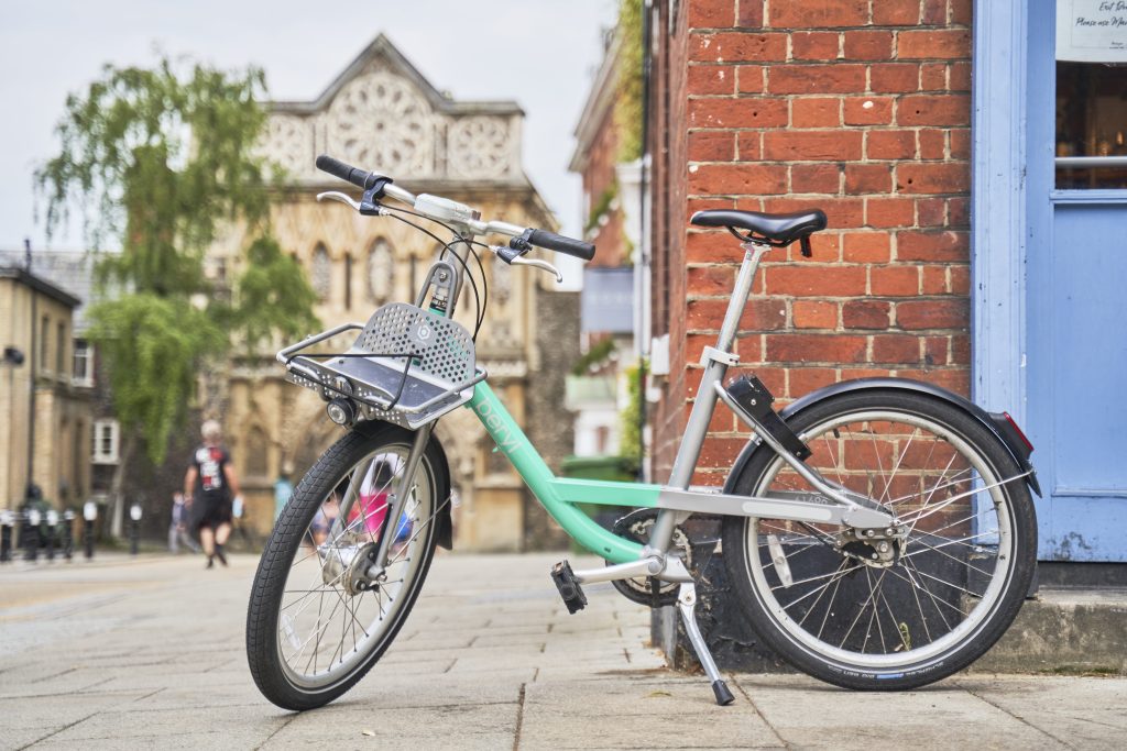 A mint green bicycle with a front basket is parked against a brick building on a cobblestone street. In the background, a blurred historic-looking structure can be seen, partially framed by trees and a cloudy sky.