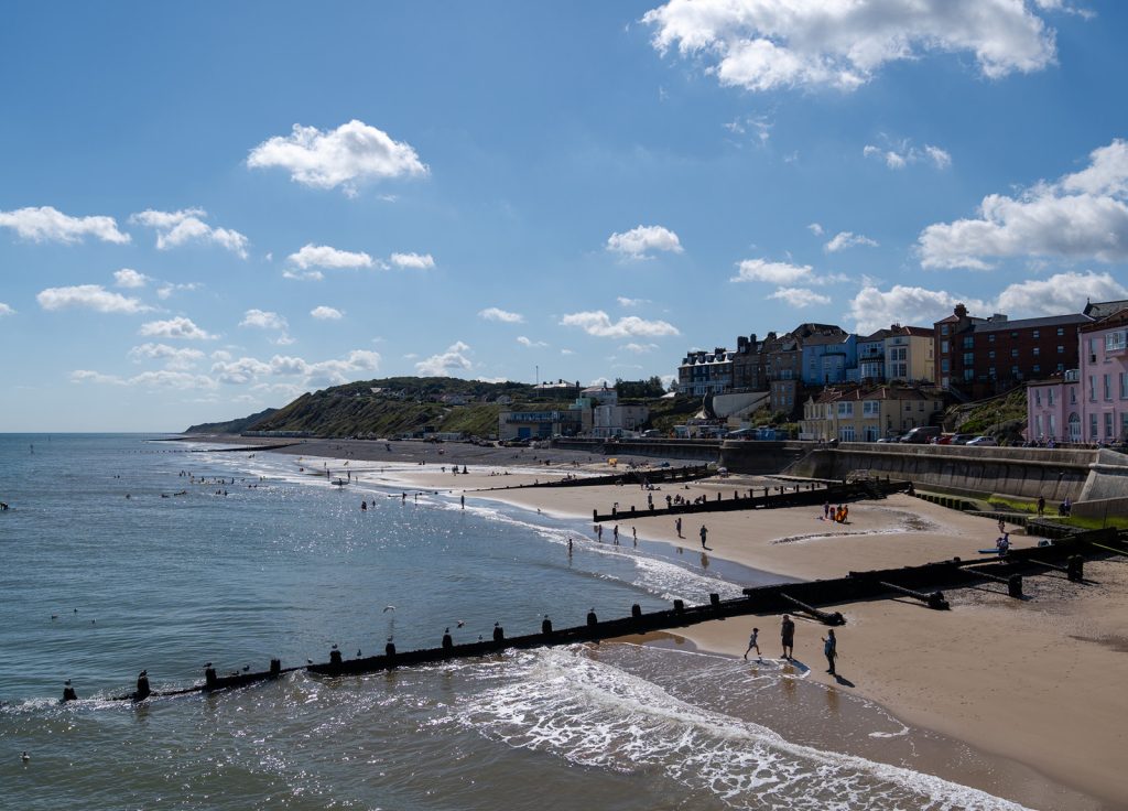 A coastal townscape with a beach featuring people walking along the shore and wooden groynes extending into the sea. The calm water reflects the sunny, partly cloudy sky. Buildings with colourful facades line the background along the coastline.