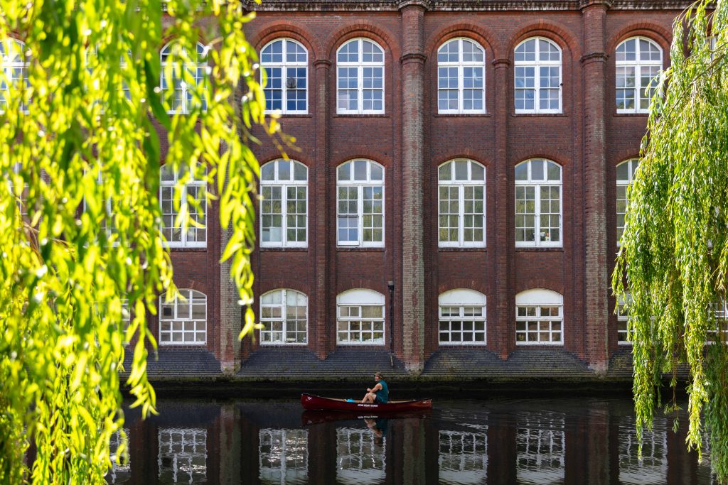 A person paddles a small red boat on a calm river in front of a large brick building with multiple arched windows. Lush green willow branches frame the scene on the left and right, creating a serene atmosphere.