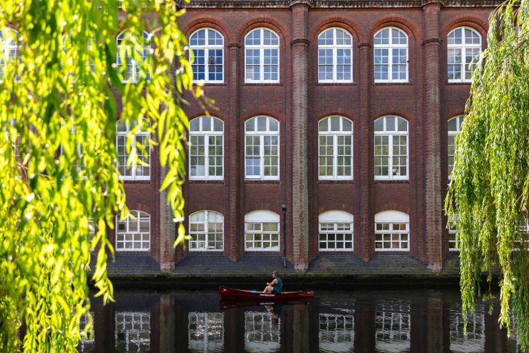 A person paddles a small red boat on a calm river in front of a large brick building with multiple arched windows. Lush green willow branches frame the scene on the left and right, creating a serene atmosphere.