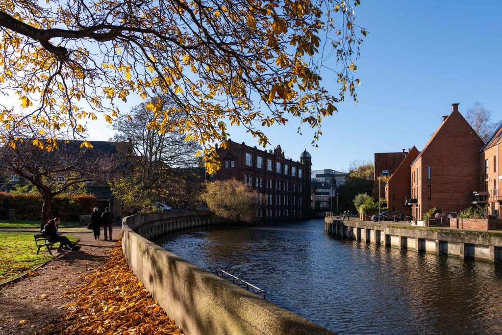 A scenic view of a river on a clear autumn day, with fallen leaves scattered on the ground and trees turning yellow. Brick buildings line the waterway, and a few people are walking and sitting on benches along the path that curves beside the river.