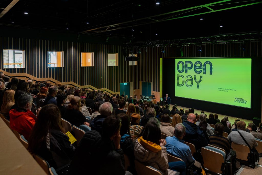 A large audience in a modern auditorium (Duke Street Riverside) watching a presentation about Open Day at Norwich University of the Arts, displayed on a large screen at the front of the hall.