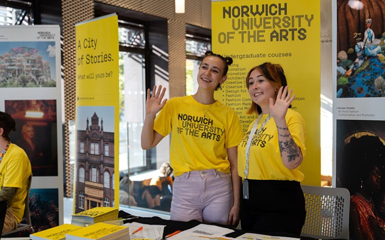 Two smiling women in yellow t-shirts stand behind a promotional booth for norwich university of the arts at an educational fair, surrounded by banners and informational brochures.